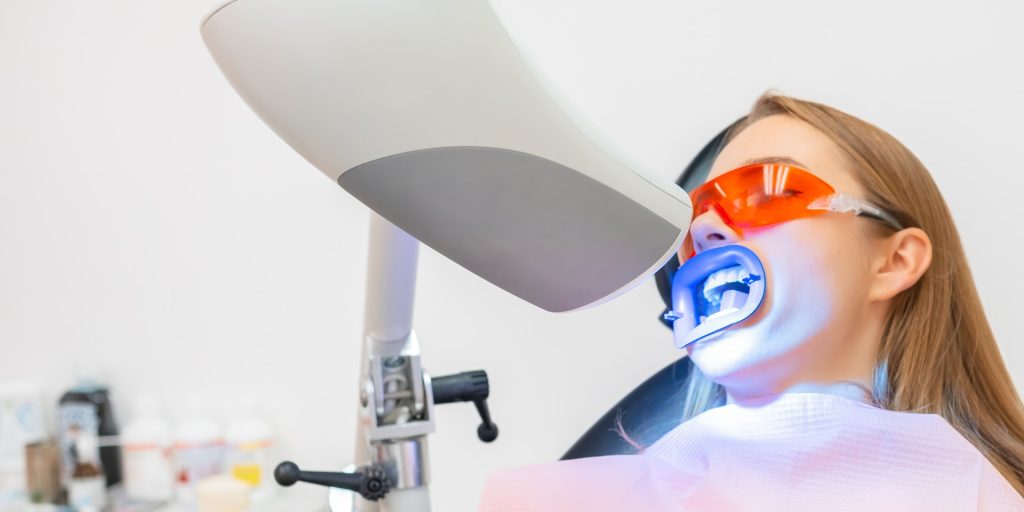 Paciente con gafas protectoras recostada en el sillón mientras la lámpara de luz azul activa el gel blanqueador en sus dientes