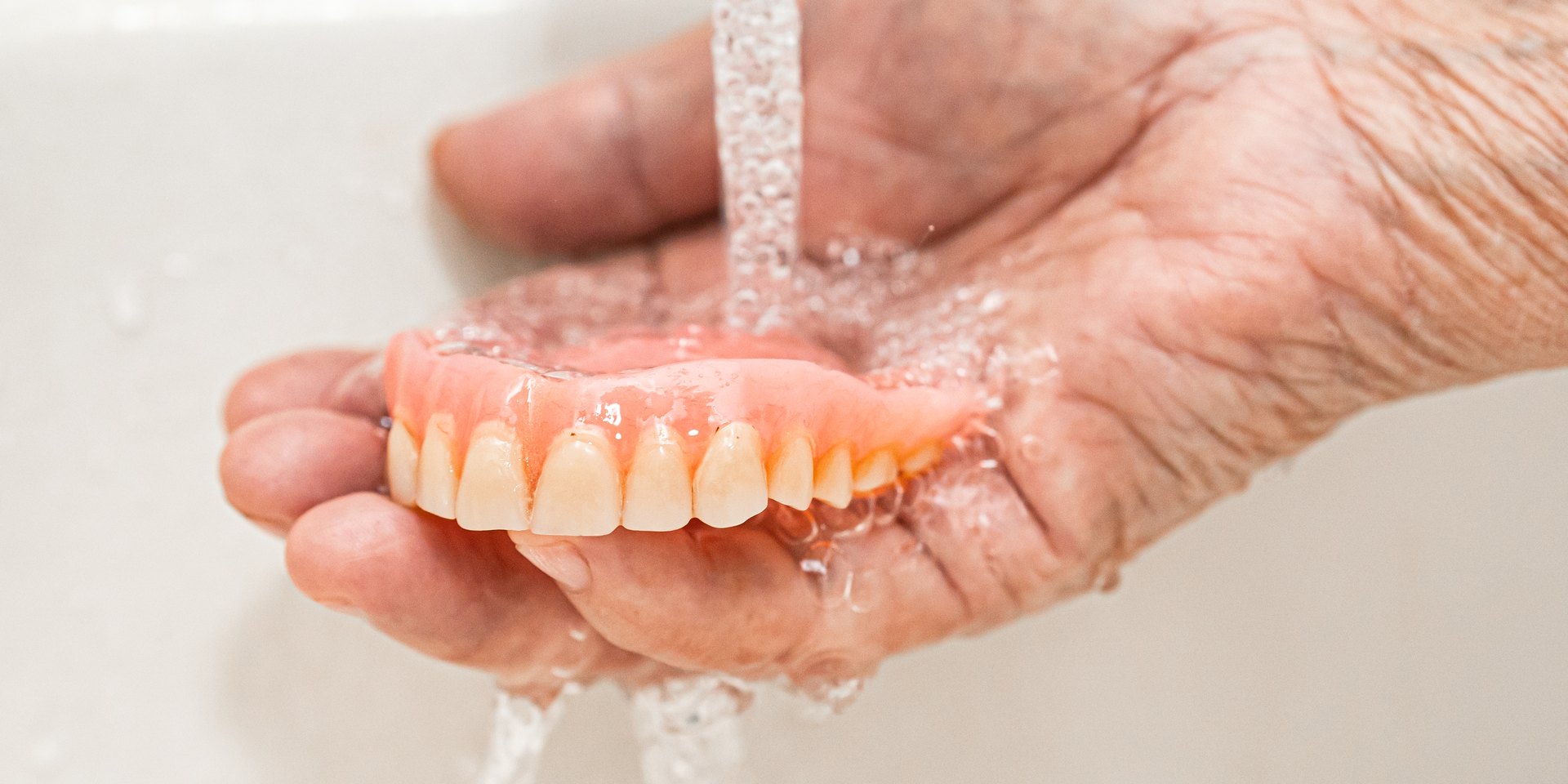 Manos de una persona mayor que aclaran una dentadura postiza superior bajo el grifo del lavabo