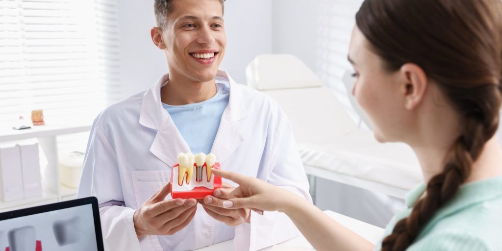 Dentista sonriente enseñando a una paciente un modelo de implante dental con corona en la consulta