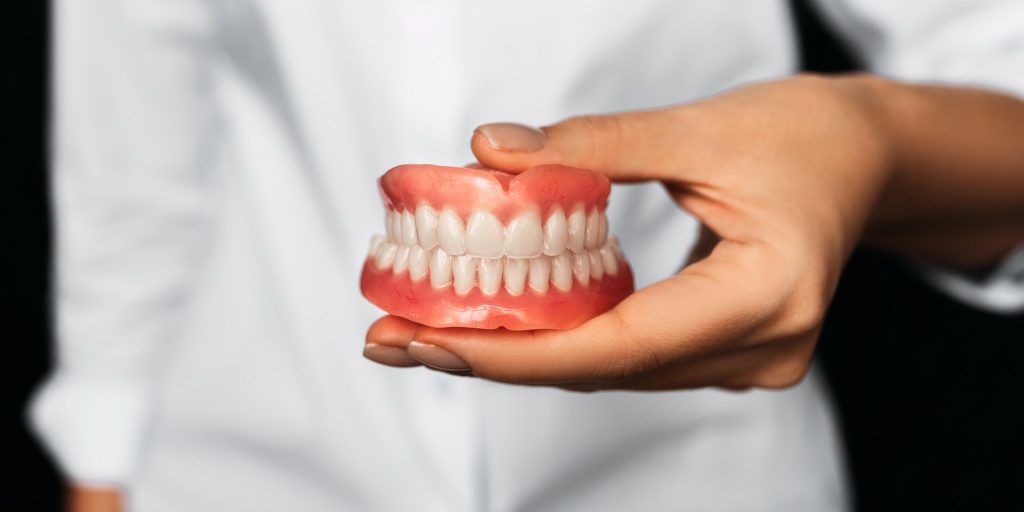 Mano sosteniendo una dentadura completa de los dos maxilares, con la encía rosada y los dientes blancos