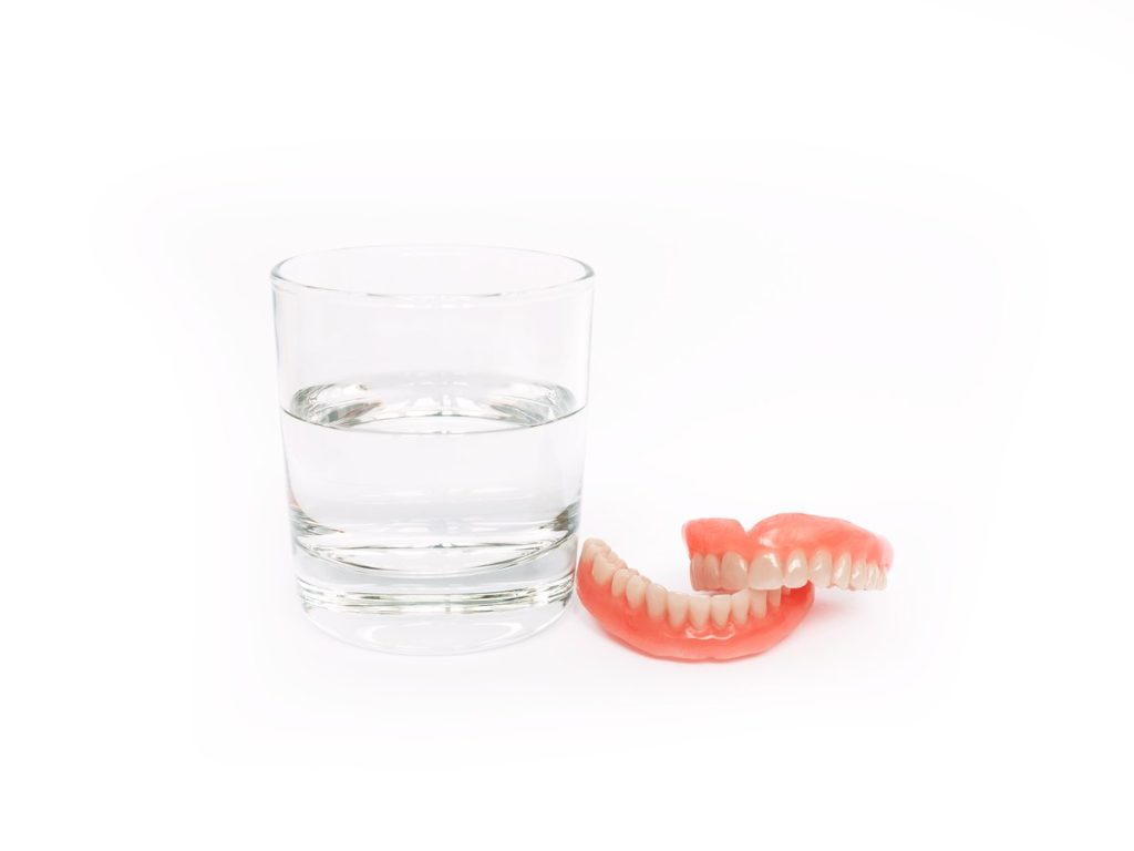 Dentadura postiza superior e inferior junto a un vaso con agua sobre fondo blanco