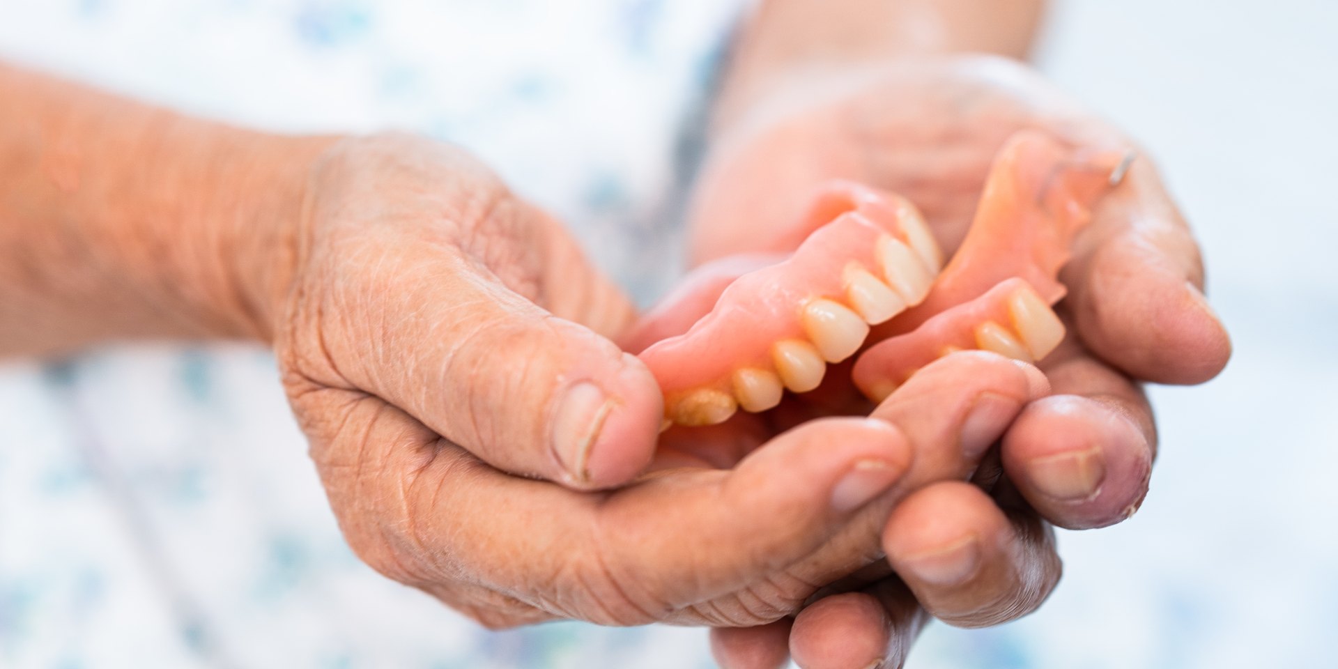 Manos de una persona mayor que sostienen dos dentaduras postizas parciales de resina, una de ellas con un pequeño gancho metálico