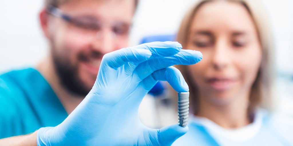 Dentista con guante azul enseñando a una paciente un implante dental de titanio sujeto entre los dedos