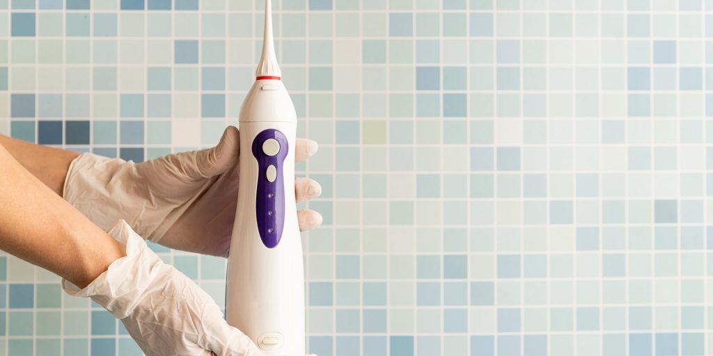 Manos con guantes sosteniendo un irrigador bucal eléctrico delante de una pared de azulejos