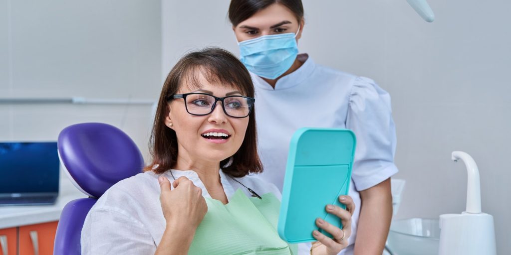 Paciente con gafas en el sillón dental que se mira al espejo con gesto alegre, junto a la dentista con mascarilla