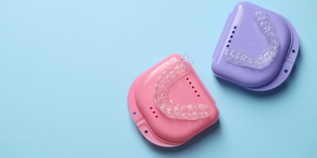 Dos retenedores dentales transparentes dentro de sus estuches rosa y lila sobre un fondo azul claro